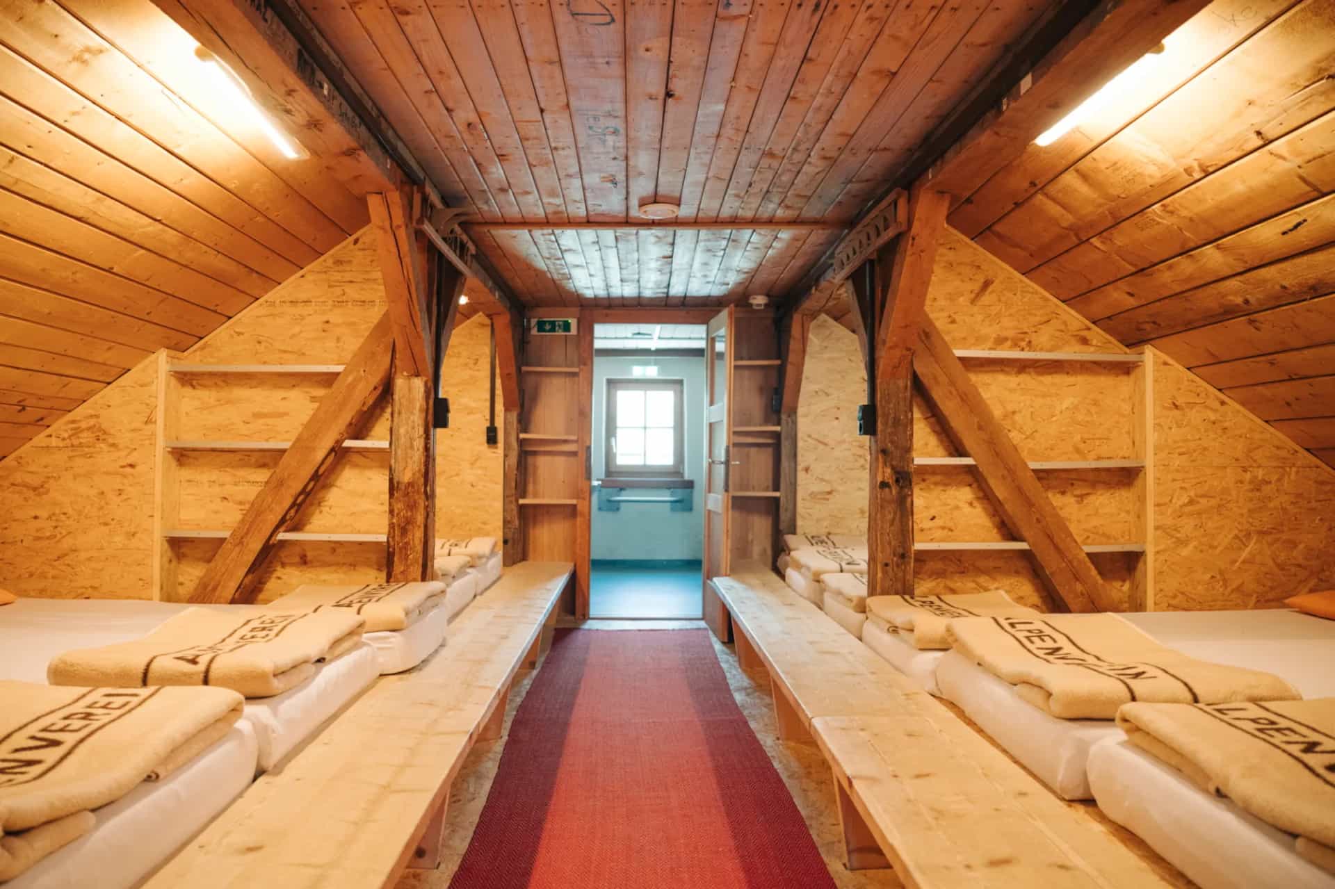 Rustic wooden dormitory with mattresses and blankets, leading to a doorway with a window.