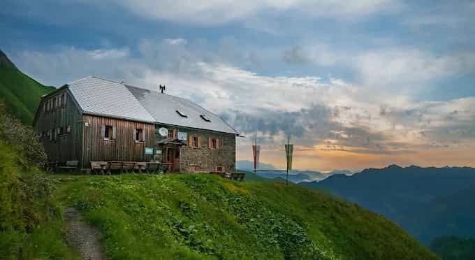 Gleiwitzer Hütte mountain refuge on grassy slope overlooking layered mountain ranges at sunset.