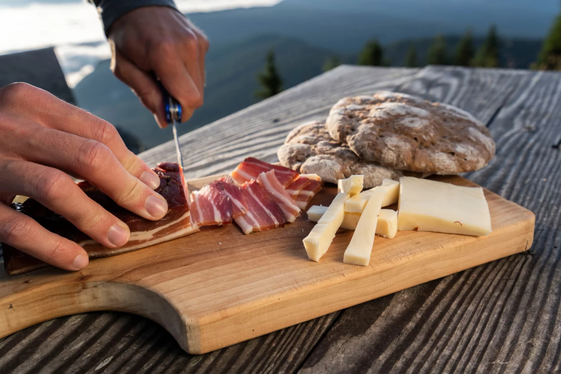Slicing smoked bacon with cheese and bread on a wooden board in the mountains