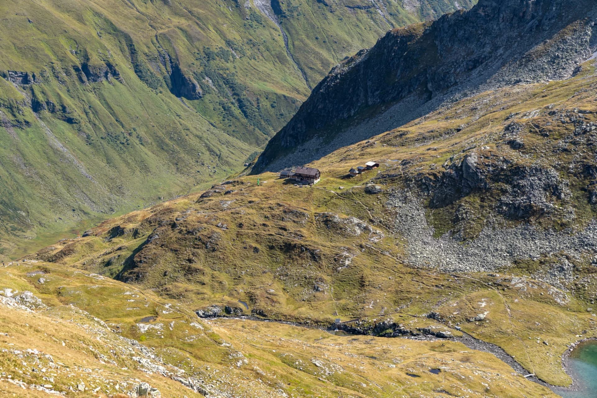 Alpine mountain hut nestled in grassy, rocky terrain with steep slopes and a small stream.