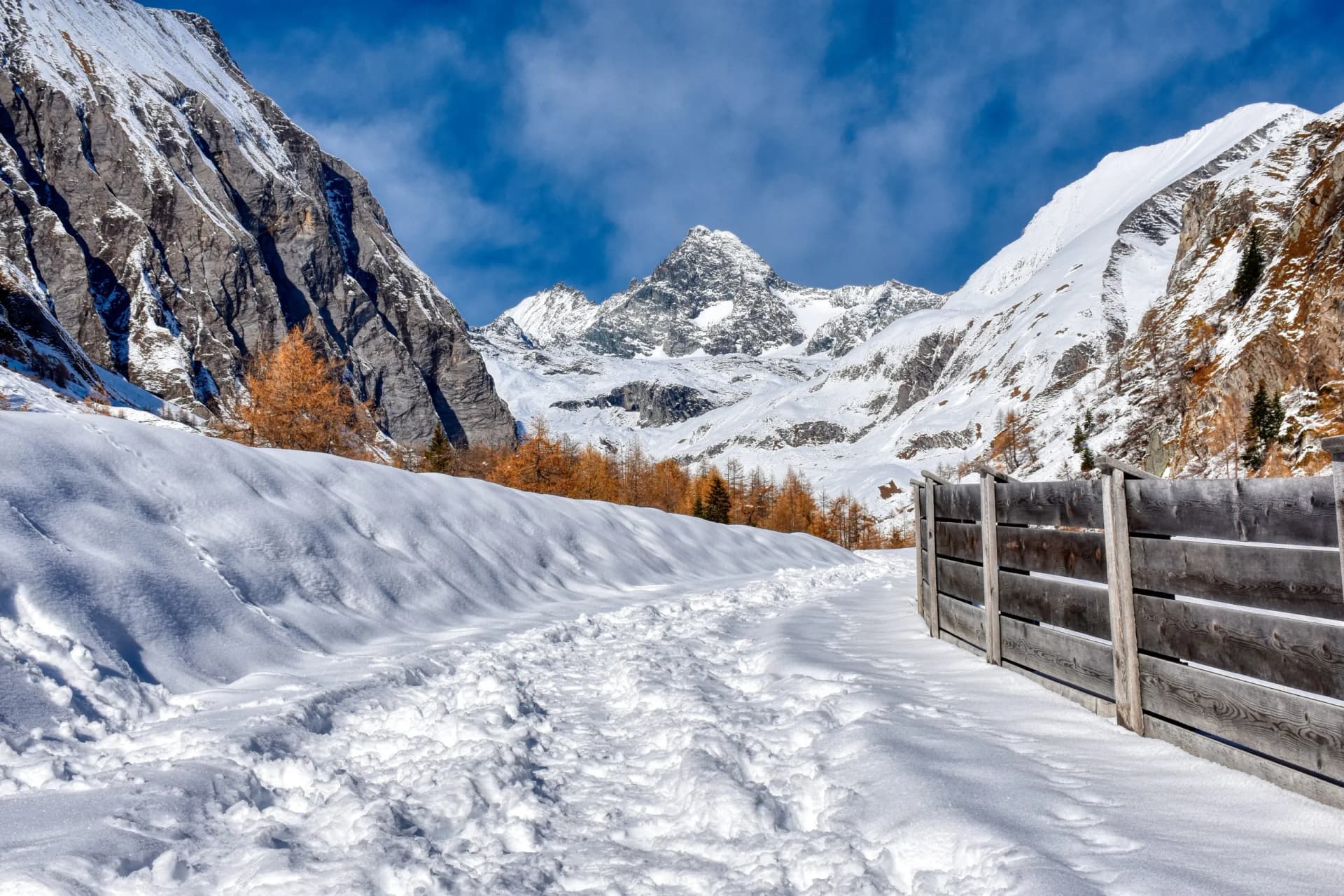 Snow-covered path leading toward snow-capped alpine mountains under a blue sky.