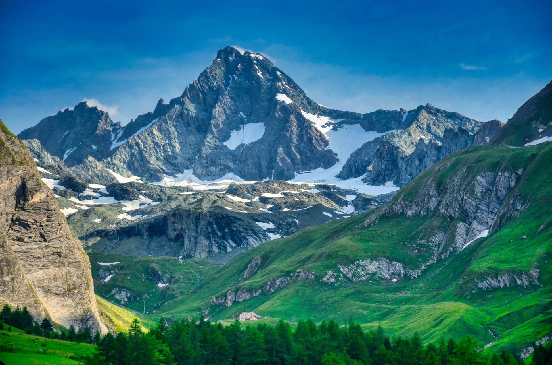 Alpine mountains with snow patches rising above green slopes and a small building, Grossglockner area.