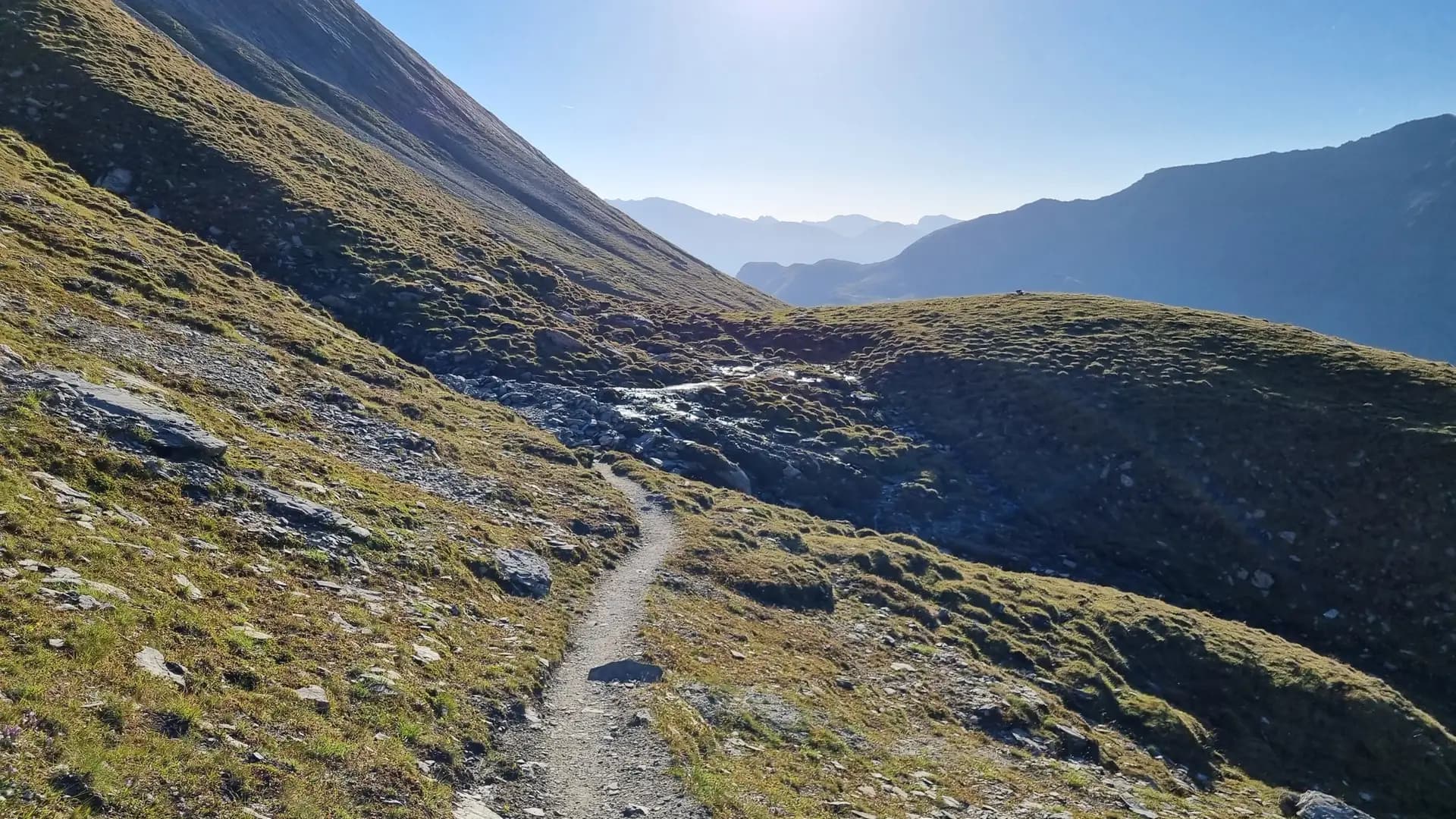 Hiking trail winding through alpine mountain landscape with a small stream and distant peaks.