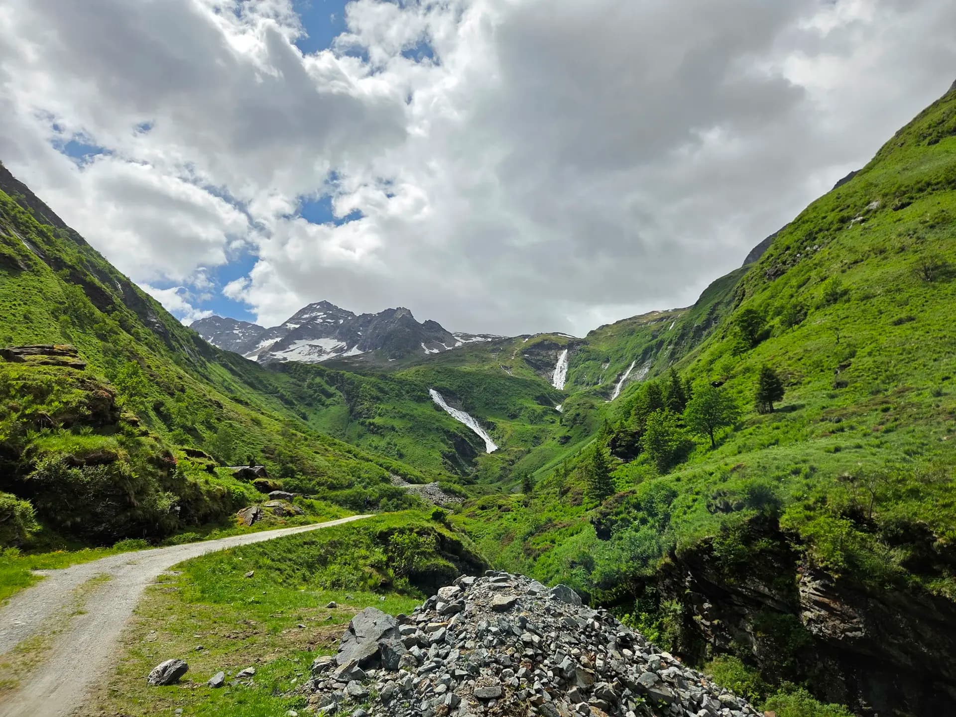 Gravel path winding through a green alpine valley with waterfalls and snow-capped peaks in the Hohe Tauern mountains.