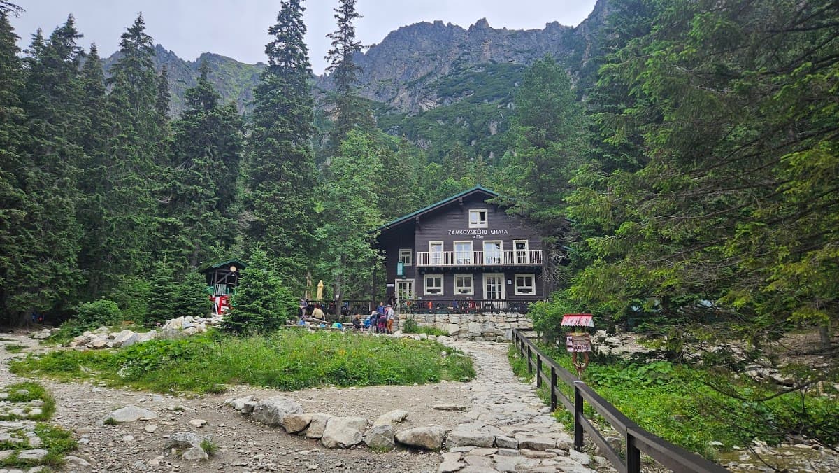 Wooden mountain chalet with people outside surrounded by pine forest and rocky peaks
