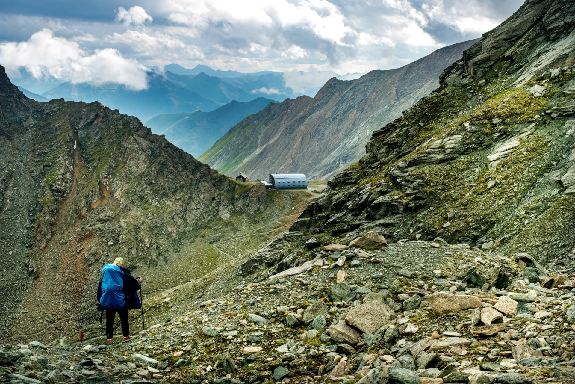 Hiker with blue backpack ascending rocky alpine terrain toward mountain hut under cloudy sky.