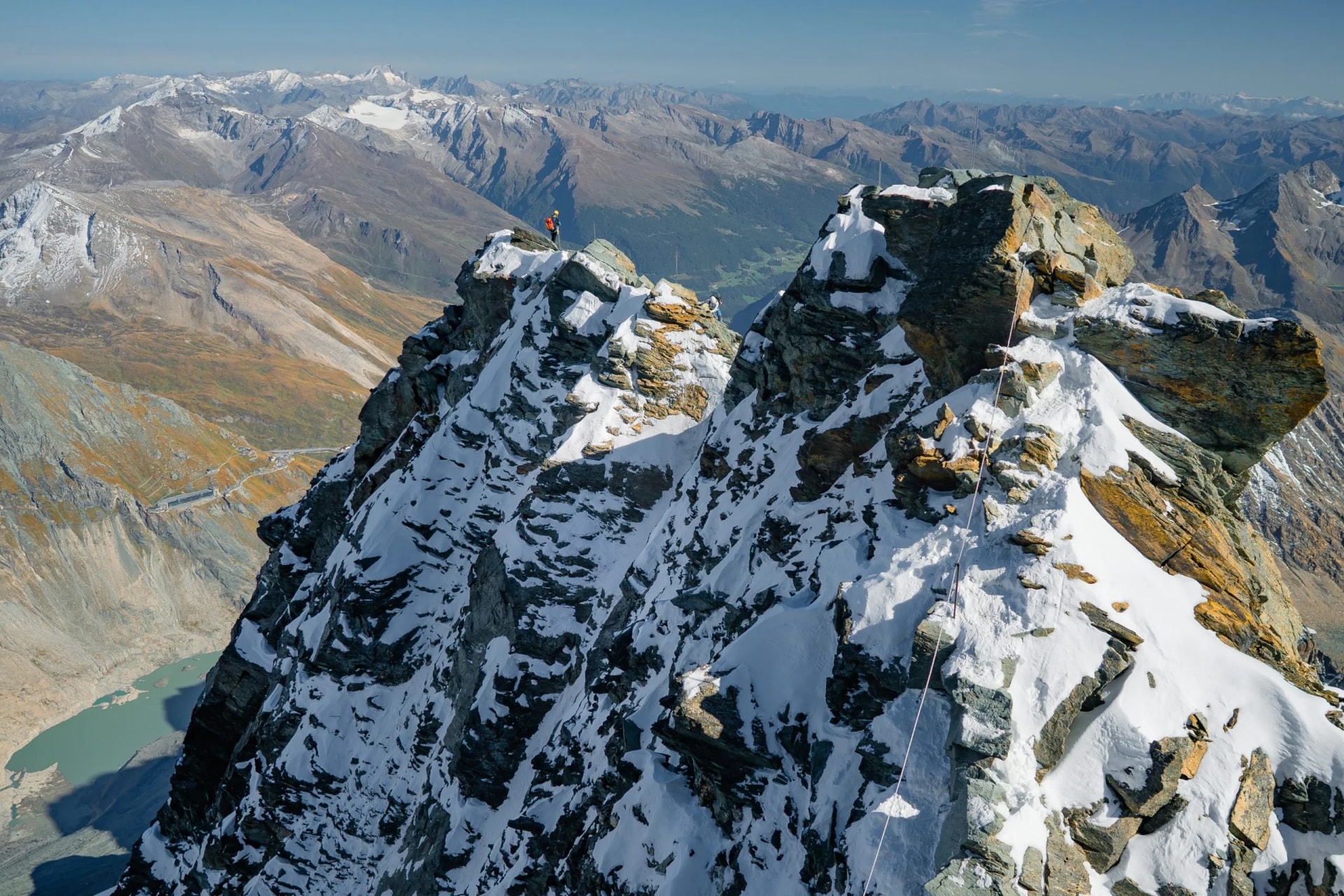 Alpinist on snowy, rocky peak with vast mountain range and lake view, Austria.