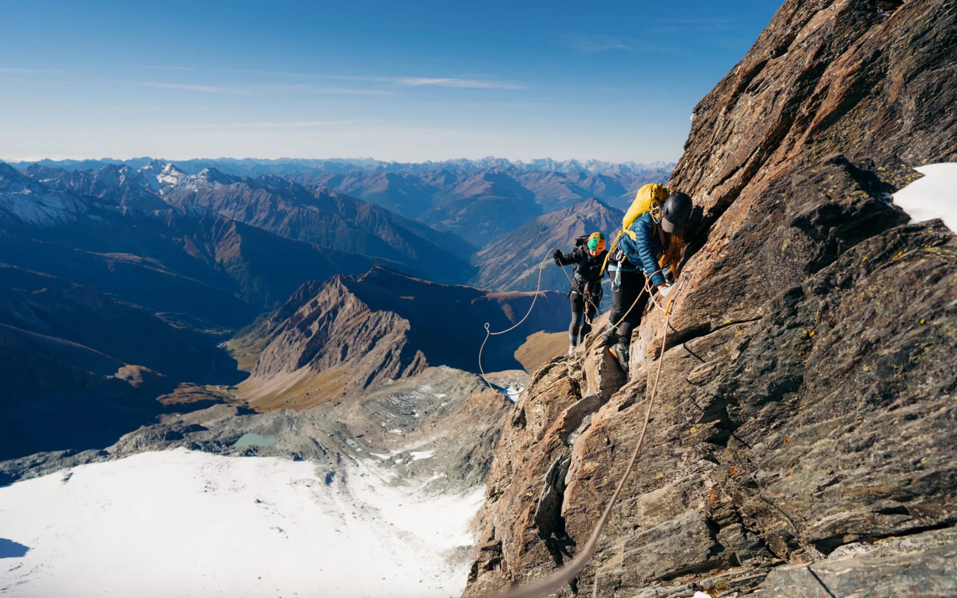 Climbers ascending a steep rock face on Grossglockner ridge above snow and alpine valleys.