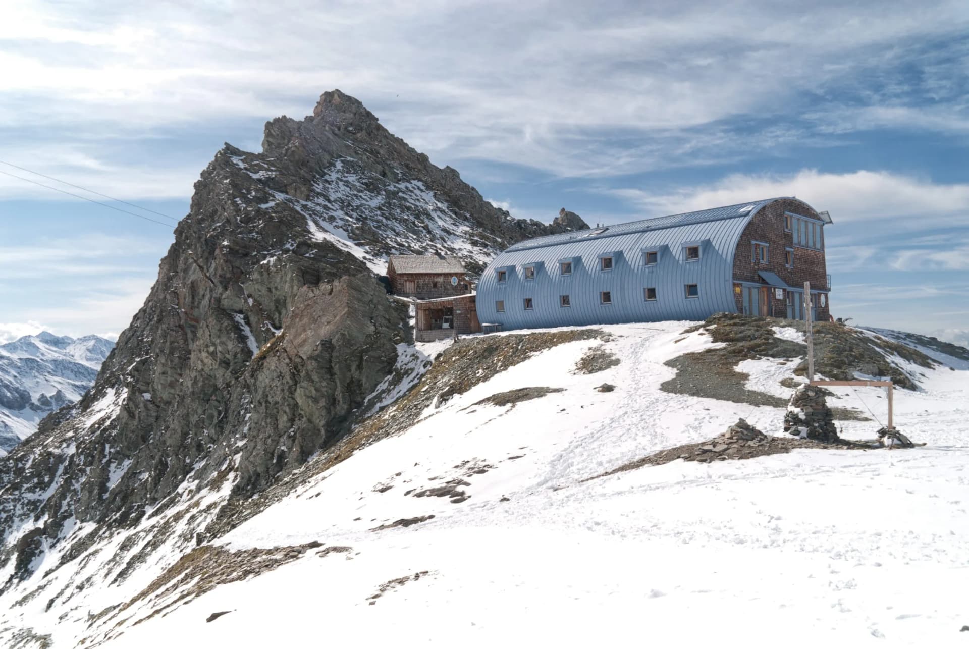 Mountain hut with blue curved roof next to rocky peak on snowy slope, with distant peaks.