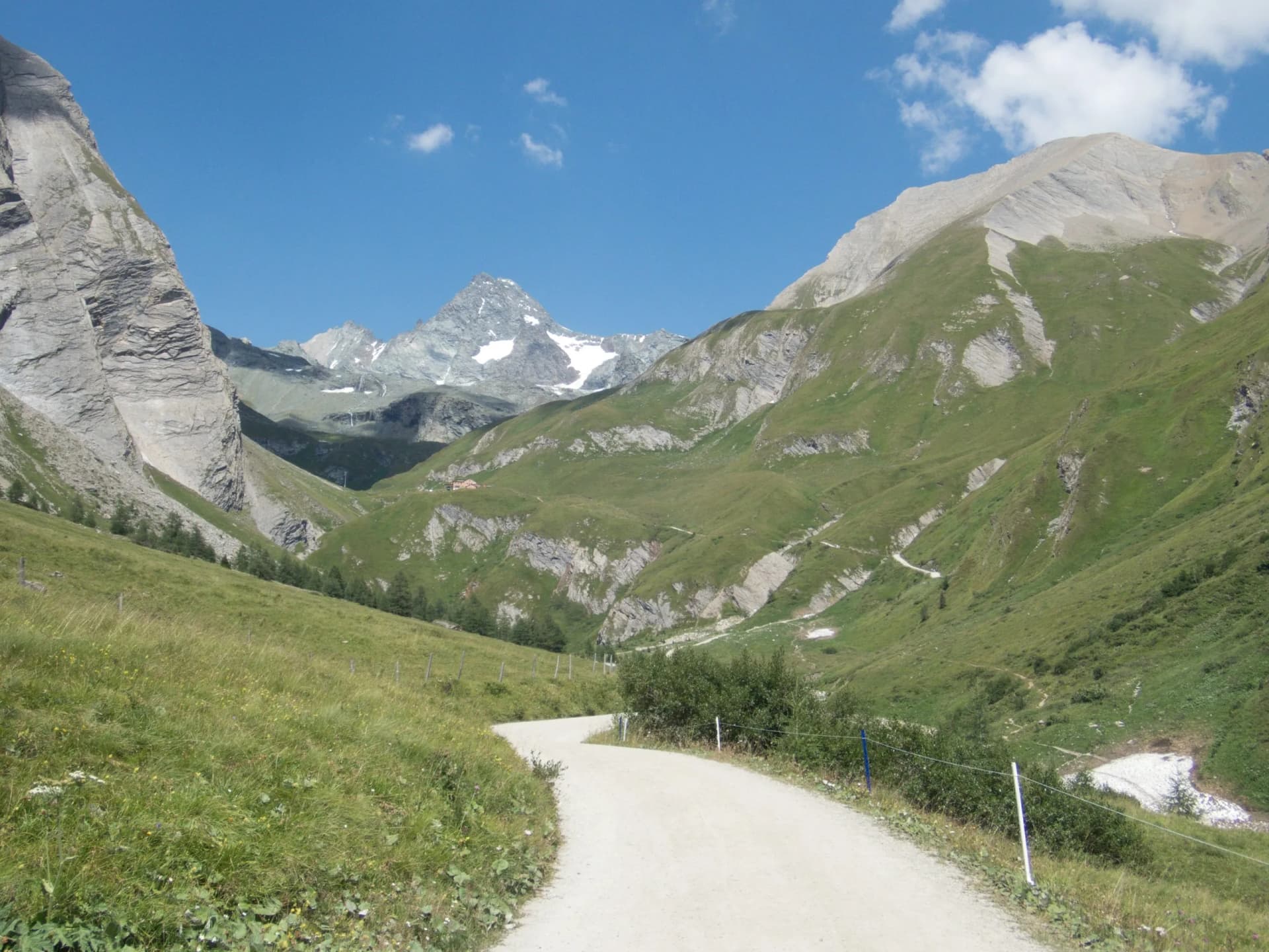 Winding dirt road through green alpine valley toward snow-capped mountains under blue sky.